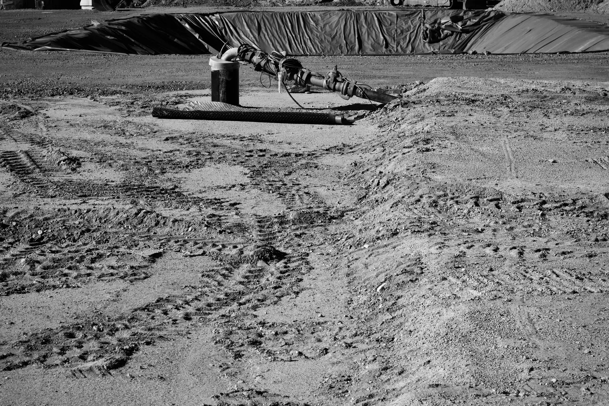 Black and white photo of tire and track marks crossing a graded site, with a pipe stack and sheeted material at the back